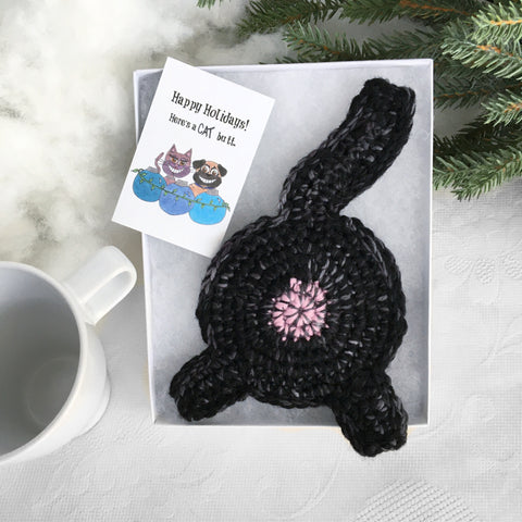 a black crocheted cat butt coaster in a white gift box with an illustrated Happy Holidays gift art card. A white mug is next to the box and it's leaning on green pine needles