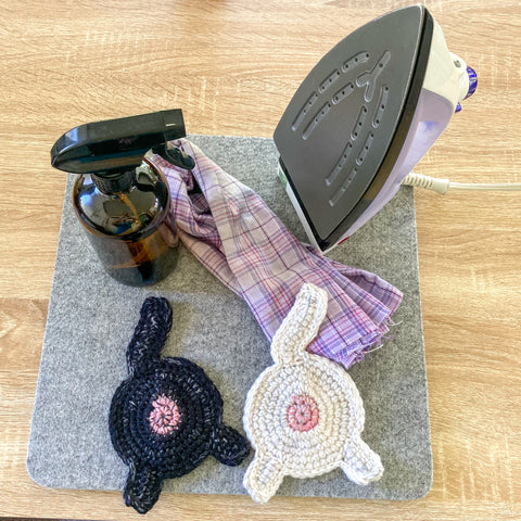 a black and white coaster each on a wool ironing surface with a brown spray bottle, a flannel piece of cloth, and an iron.
