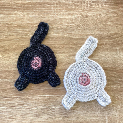 a black and a white crocheted cat butt coaster on a wood surface