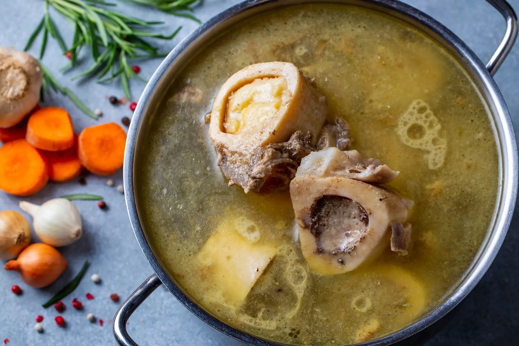 A pot of simmering bone broth with large marrow bones is surrounded by fresh carrots, onions, garlic, herbs, and peppercorns.