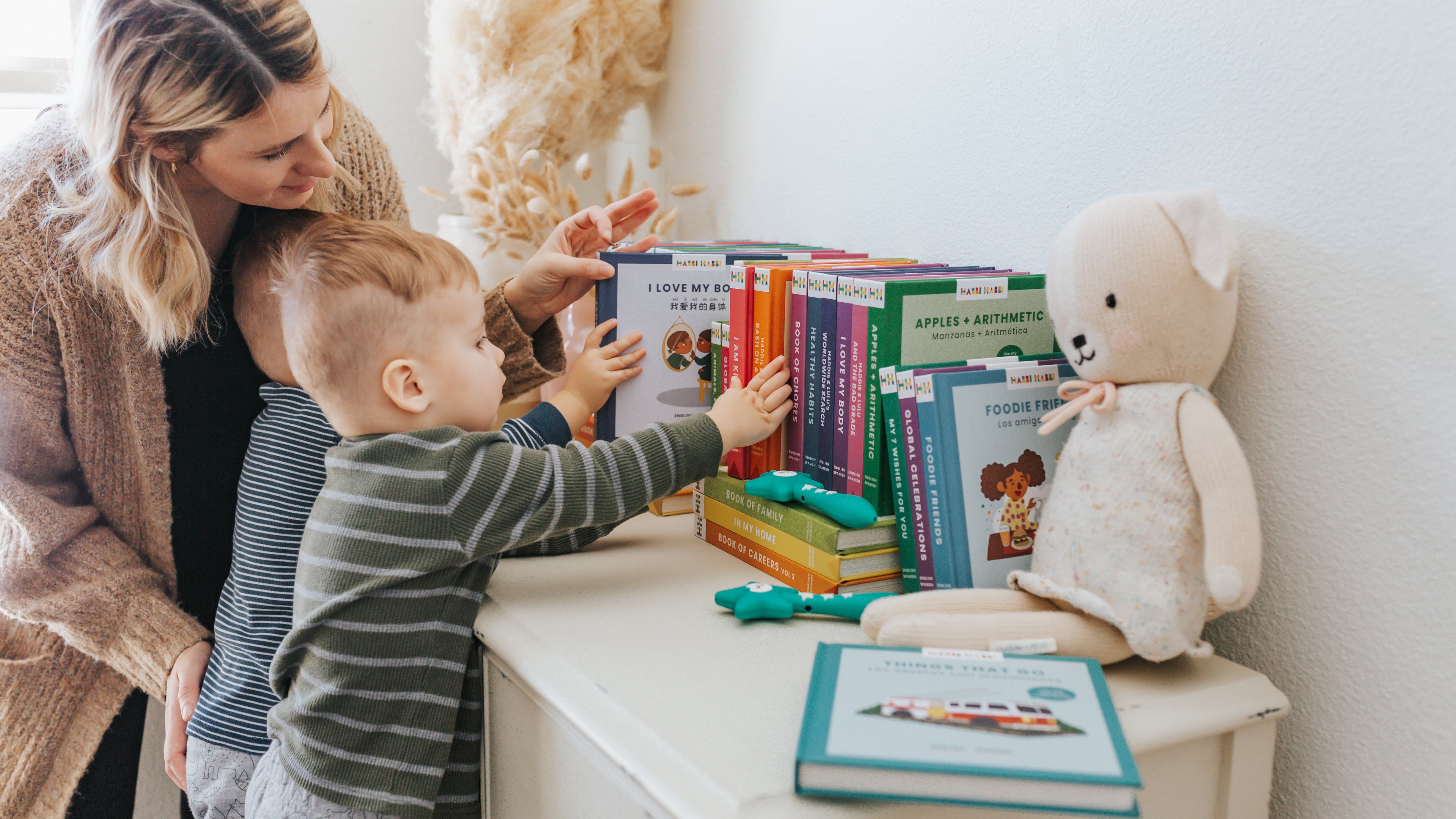 Mother selecting a Habbi Habbi book from a shelf with two toddler-aged boys.