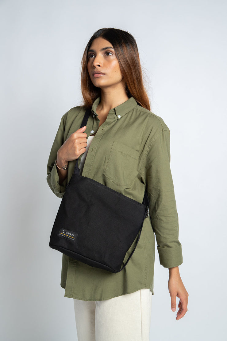 Picture of woman wearing a Trakke Kelso Sling Bag in Black. Grab handle at right side. 5L, 5 litre capacity. Dry-finish waxed canvas everyday bag with black cotton webbing.
