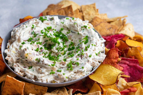 A bowl of bacon and onion dip topped with chopped chives on top of a platter of multicolored vegetable chips