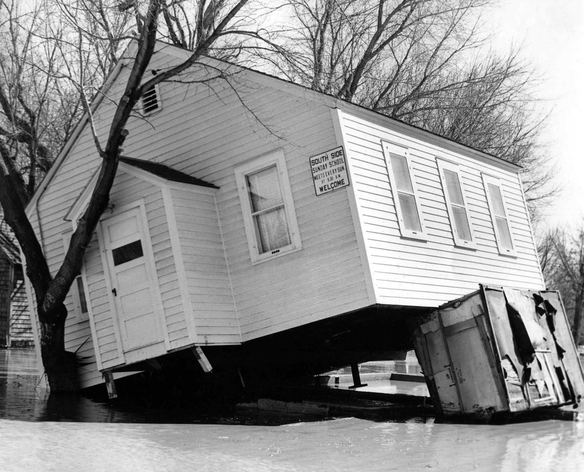 Sturdy trees saved the South Side Mission from being washed away downstream, Bismarck, April, 1952. Located on the corner of Sweet Avenue and Washington Street, the little church stood fast against the flood for several hours. Finally it was unable to withstand the rushing waters and was tilted up on its side against the trees. -- BISMARCK TRIBUNE