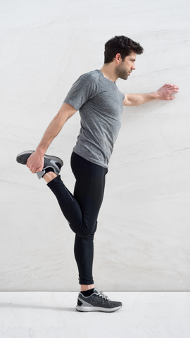 man in bright room wearing grey shirt and black leggings. He has one hand on the wall and the other holding his foot behind him in a standing quad stretch