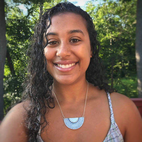 Young brown woman with curly hair smiling, with a silver half moon necklace (Deep Knowing Necklace), wearing a dress with straps. Green trees are behind her. 