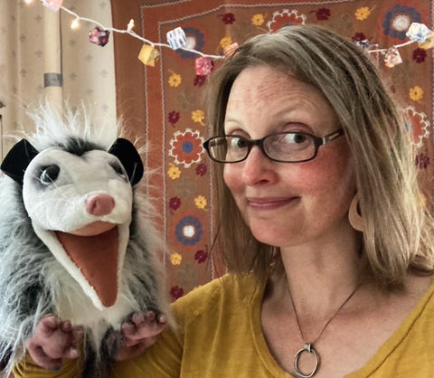 Ann Braden, a white woman with blond hair and glasses, smiling, wearing the Zen Coil Necklace. She's holding a puppet of an opossum