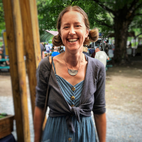 Picture of a white woman in her 40s smiling at a farmers market, wearing the Deep Knowing Necklace and a blue dress. It's summer and she has pigtails.
