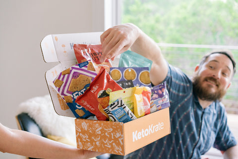 smiling happy guy in a blue shirt reaches into a KetoKrate to grab a keto friendly snacks.