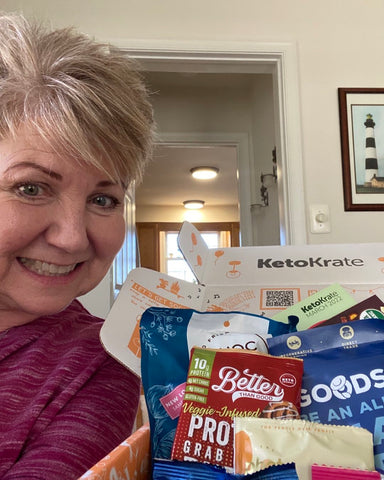 successful, middle aged women in a burgundy shirt posing with her keto friendly snacks from KetoKrate. Another happy member!