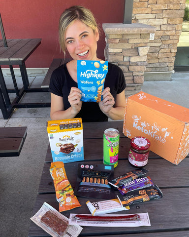 Smiling blonde woman sitting at a picnic table holding snacks from her KetoKrate