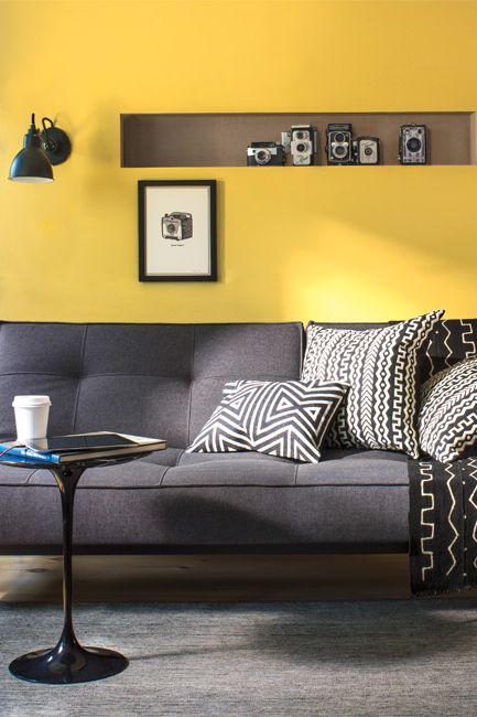 An armless gray couch and small, circular coffee table set against a yellow accent wall features black and white patterned pillows.