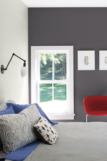 A bedroom in varied gray tones features a contemporary red accent chair and two windows with a view of outdoor greenery.