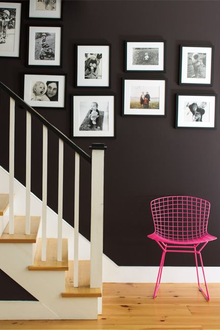 A staircase with banister is complemented by black and white family photos on the wall with a hot pink wire chair on the landing.