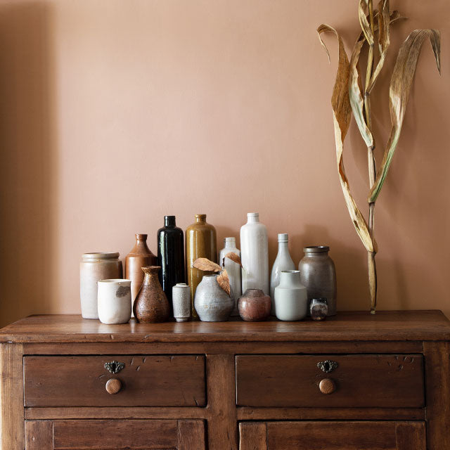 A collection of bottles on a wooden credenza in front of a gold-painted wall, with dried plant décor.
