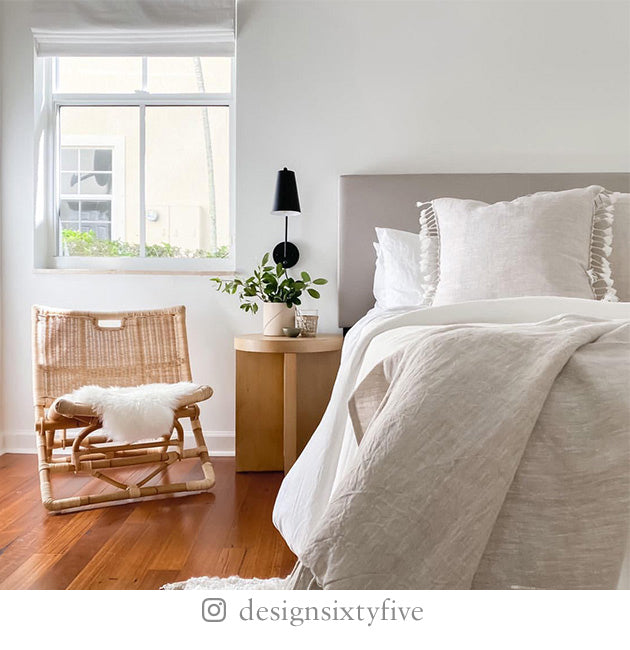 White-painted bedroom with gray bed linens, tasseled pillows, rattan chair with white fur throw, and wooden end table.