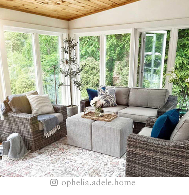 Sunroom with large windows, loveseat and two chairs, ottomans, rug, and various plants, surrounded by trees.
