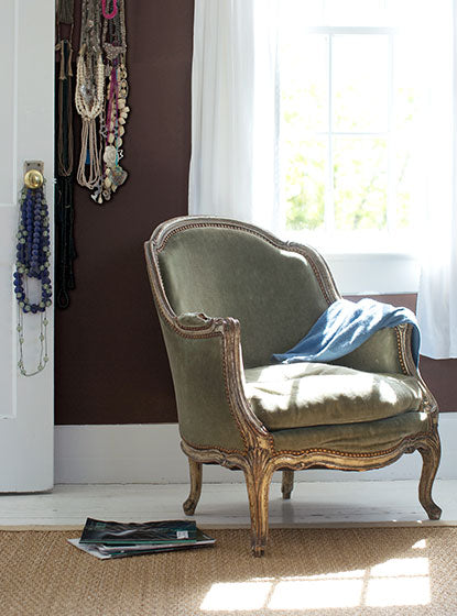A gray vintage-style chair in a brown-painted room with various necklaces on the wall and hung on a doorknob on a white door.