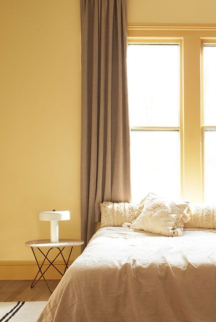 A gold-painted bedroom with a bed on a striped rug in front of a large window with beige curtains, featuring an end table and lamp.