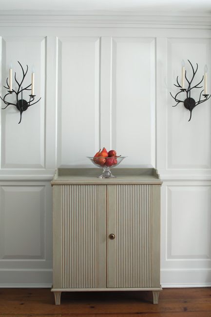 Panelled gray-painted walls frame a chest centered between candle sconces.