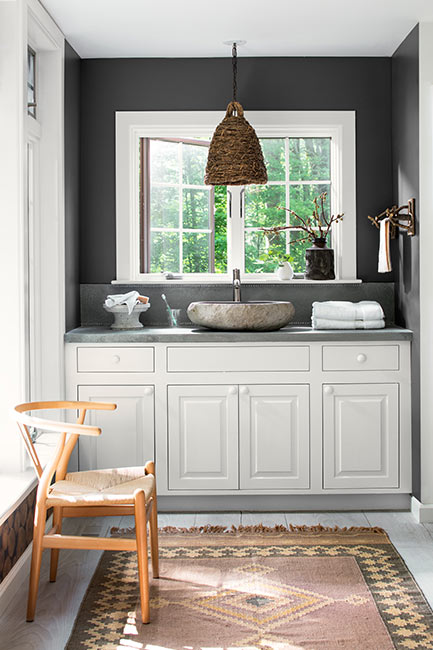 A white bathroom vanity with stone vessel sink underneath a charcoal gray wall with window.