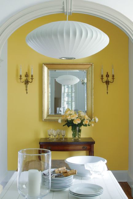 A dining room features a contemporary lamp at center, a contrast to the traditional furnishings including a white wooden farmhouse table, antique white chair, ornate sconces and arched windows seen in the neighboring hallway.