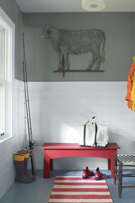 A mudroom features a large illustration of a sheep against a gray-hued painted wall panel; a red bench and striped red and white rug add a punch of color in contrast to white-tiled walls.