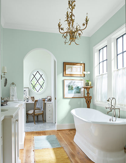 Airy bathroom with pastel green walls, chandelier, soaking tub, sink and cabinets, wall art, large windows, and a desk and chair.