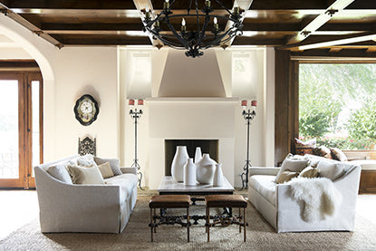 Large open living room with coffered ceiling, chandelier, white seating, leather stools, candelabras, and large windows.