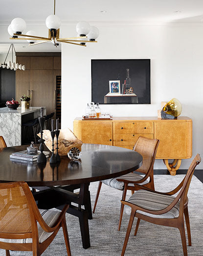 A white modern dining room with a light hanging above a large black round table and brown chairs next to artwork hung above a long console table.