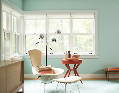 Corner of a room with blue-green walls and large windows, including a sitting chair, stool, and side table.