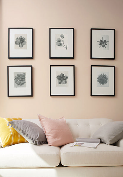Dusty pink-painted walls with six picture frames hung above a white couch with yellow, pink, and gray throw pillows.