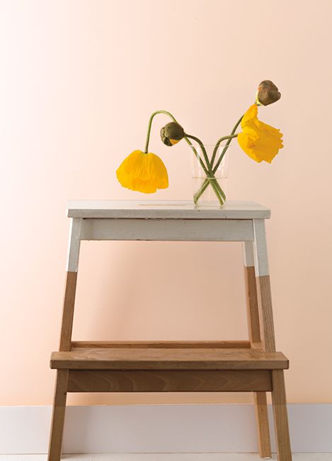 Pale peach walls with two yellow flowers in a vase set on top of a two-toned step stool.