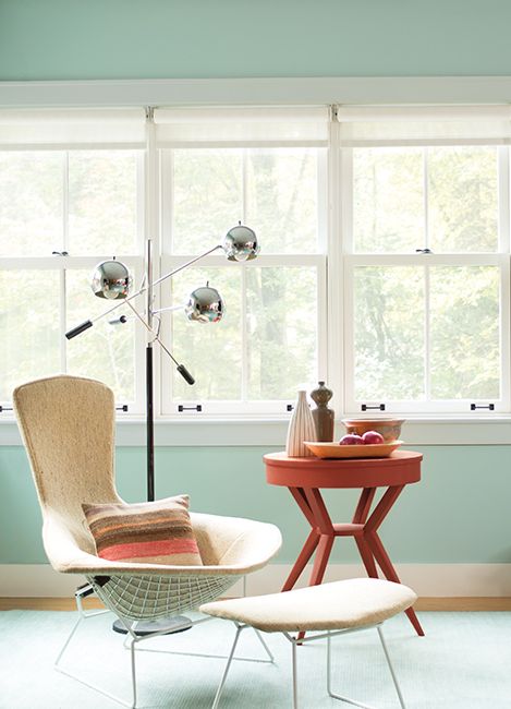 Corner of a room with blue-green walls and large windows, including a sitting chair, stool, and side table.