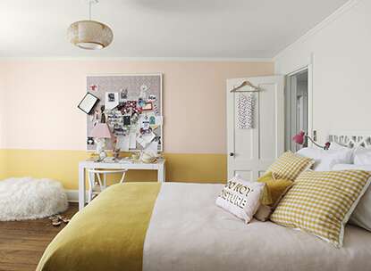 Soft white- and yellow-painted bedroom with beanbag chair, cluttered corkboard, white desk, and bedspread with two-toned bedding.