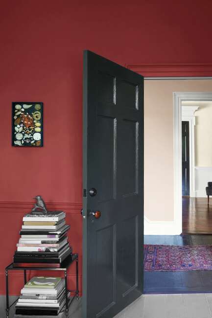 A Pomegranate-painted wall with Ebony King-painted door, stack of books on an end table and wall art, leading to a hallway in Head Over Heels.