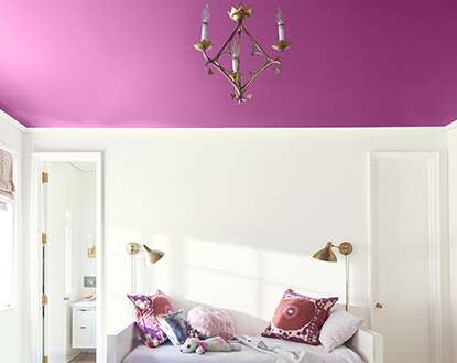 White bedroom with white bed and matching bedding, multicolored pillows, geometrical end table with books and a stuffed dog.