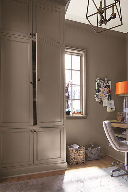 A taupe-painted home office with cupboard, beige chair, wooden desk, note board, white ceiling, and abstract chandelier.