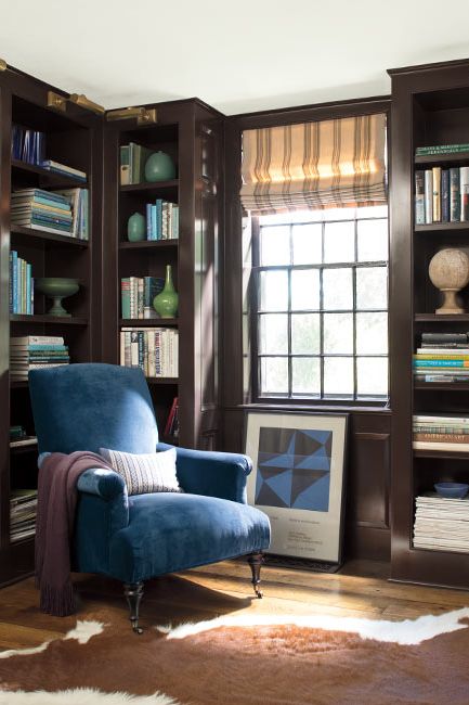 A deep blue-painted library with built in shelves, an animal hide rug, and dark blue velvet chair.