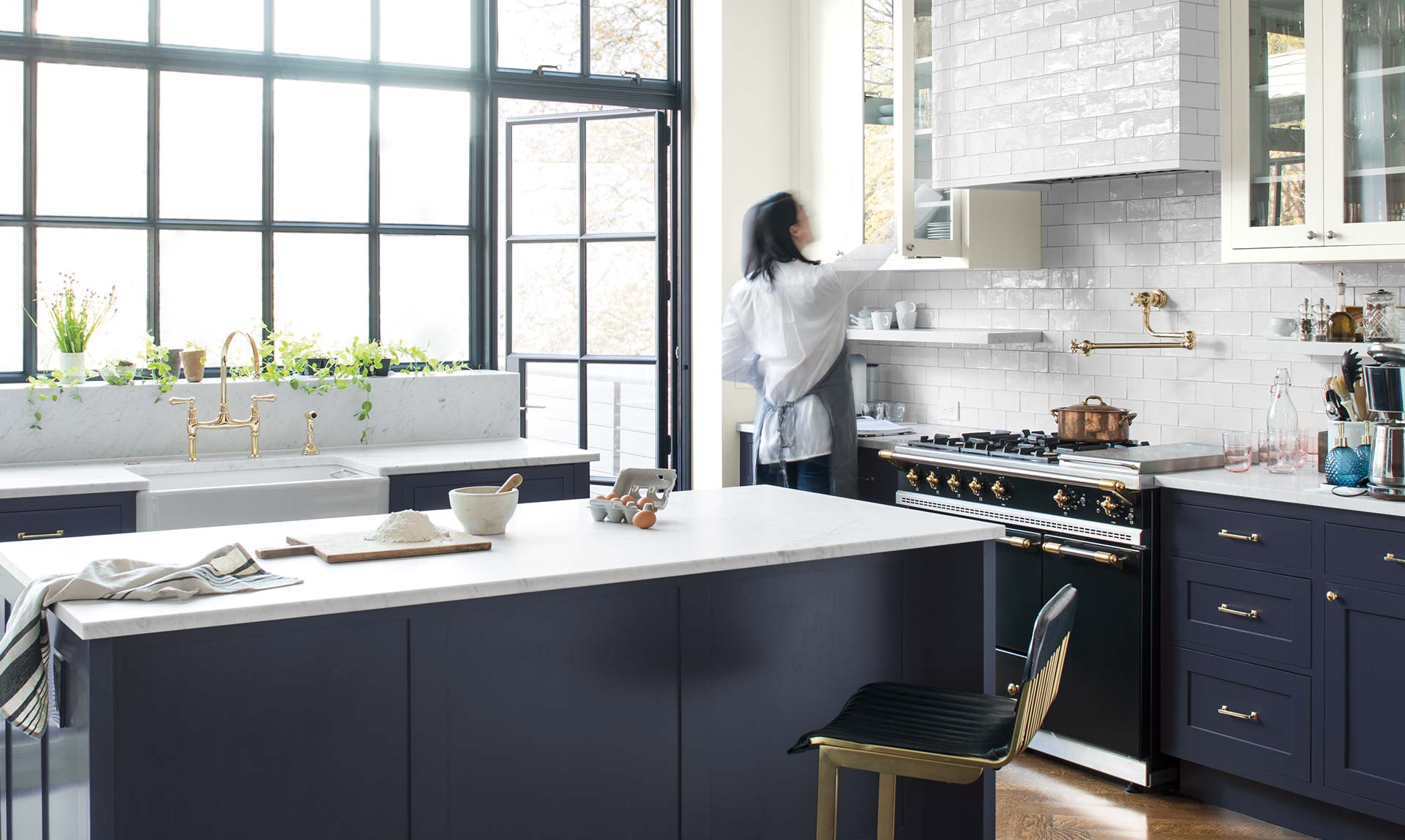 A woman reaches into a white-painted cabinet in an airy kitchen with a navy island. Color of the Year 2019