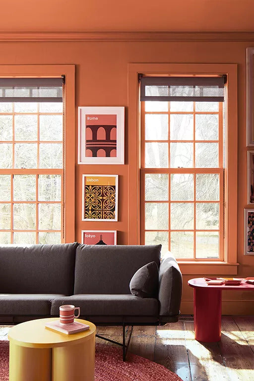 A bold living room with walls and trim painted in a deep radiant orange includes travel wall posters, a dark gray couch, round yellow coffee table, and two over-sized paned windows.
