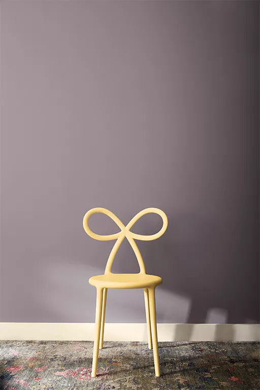 A small chair with a painted light yellow chair with bow-shaped back in front of a midtone violet wall with white baseboard and multi-colored area rug.