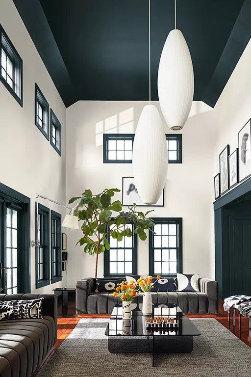 A dramatic great room with a high ceiling and window trim painted in a deep green, off-white-painted walls, large charcoal gray couches, a black lacquer cocktail table, and oversized oval pendant lighting fixtures.