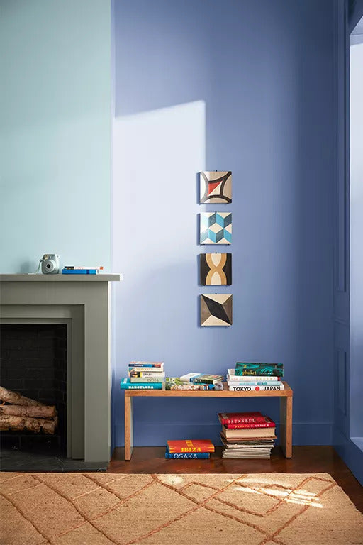 The corner of a living room with a wall painted in a pale crisp blue, a fireplace, orange area rug, and a book-filled bench beneath four small art pieces.