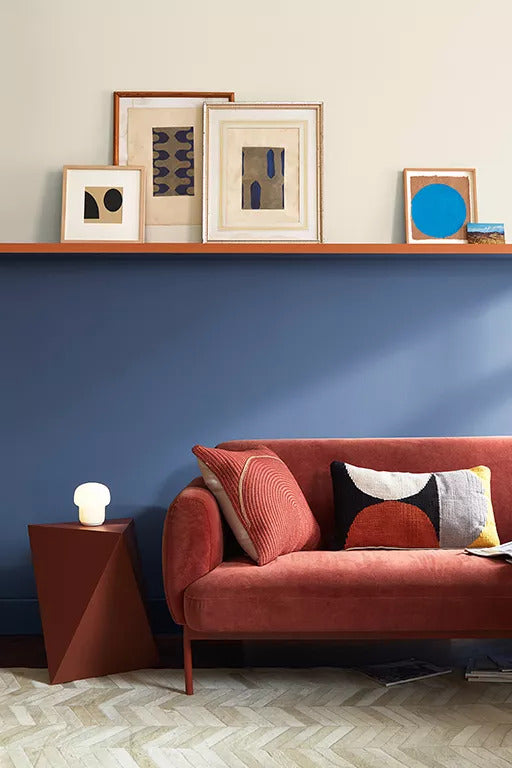 A living room with a split wall separated by a shelf painted in a sumptuous blue with hints of violet on the lower half and an off-white hue on the upper wall, a shelf holding several frames, and an orange velvet couch with graphic pillows.