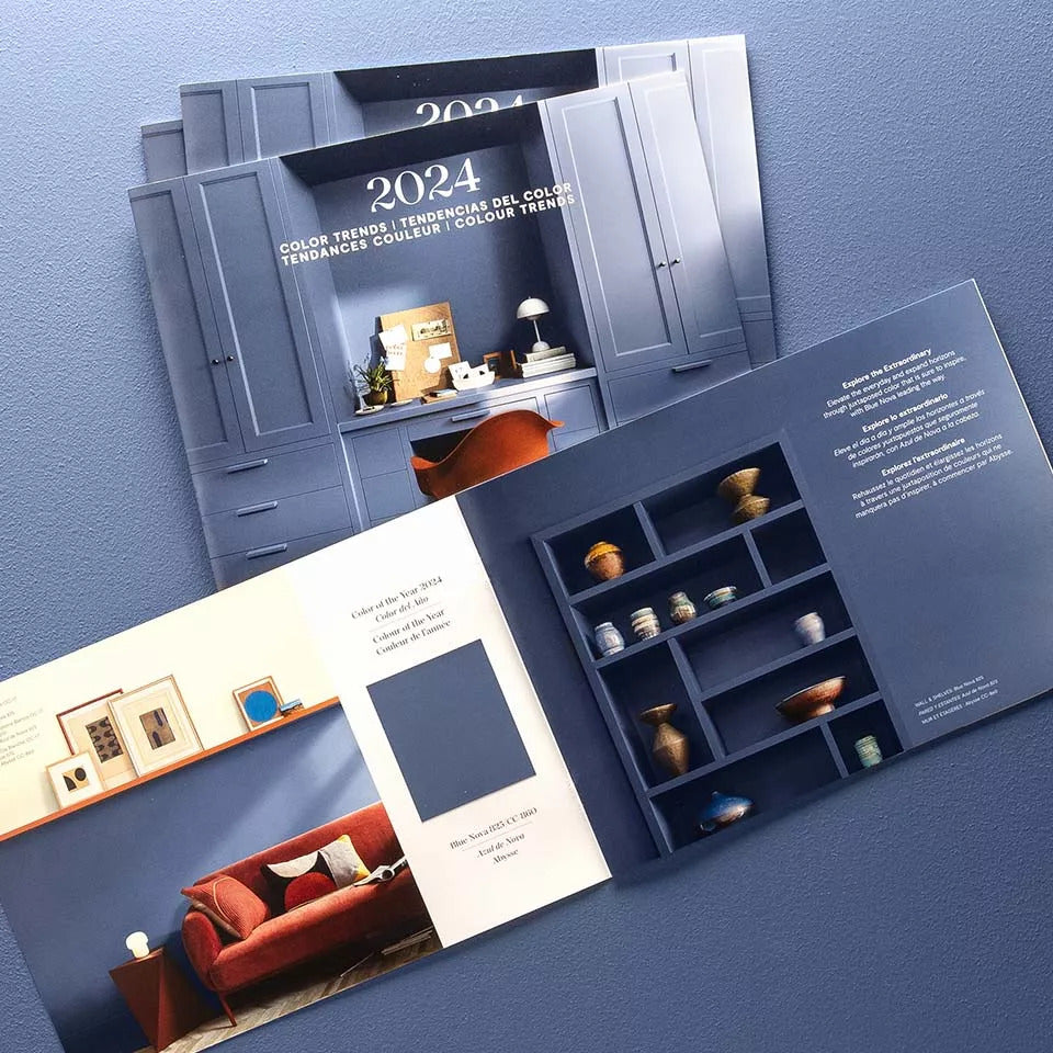 A stack of Benjamin Moore Color of the Year 2024 brochures, with the one on top open to a middle page showing a Blue Nova paint chip.