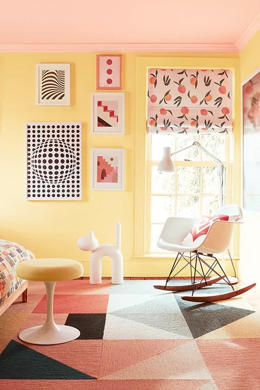 A playful painted bedroom in vibrant tones including a soft pink-painted ceiling and light yellow-painted walls, a rocking chair, peach-pattered blinds and a range of graphically bold and colorful pillows and art.