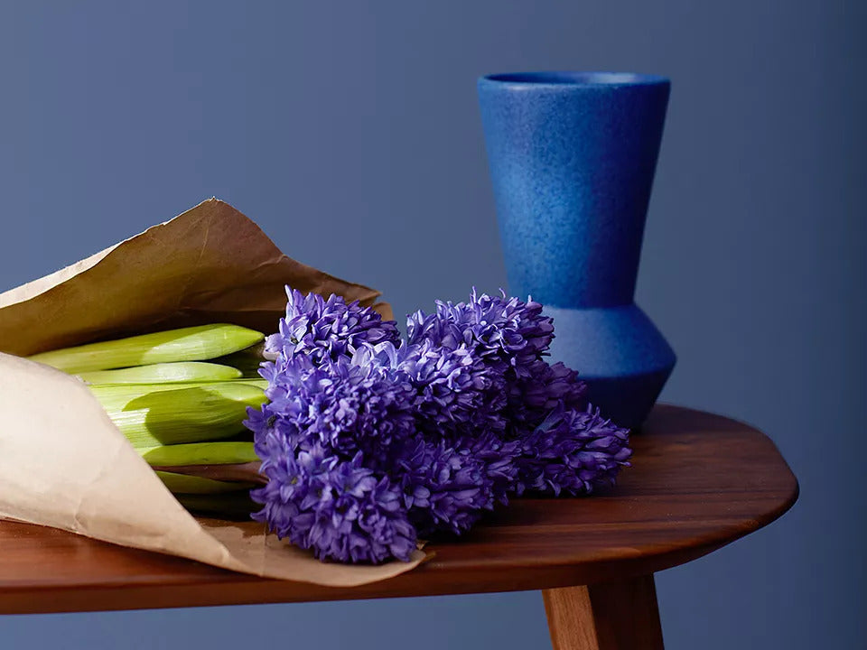 A deep blue wall with decorative trim behind a small wooden table holding a cobalt vase and a bouquet of purple flowers wrapped in brown paper.