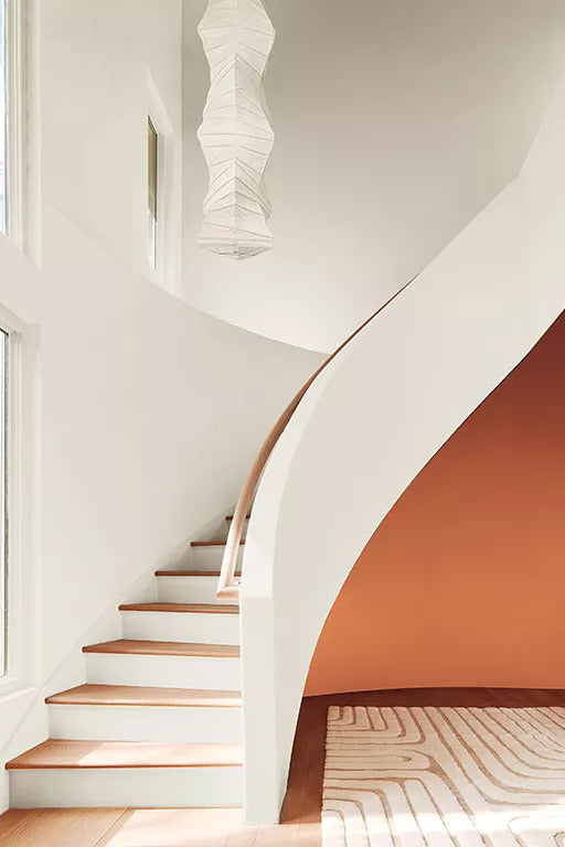An off-white-painted curved staircase a bold red accent wall beneath it, neutral area rug, bright natural light streaming through large windows and a rice paper chandelier.