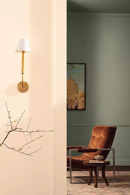 A rosy, off-white-painted hallway with a brass sconce and decorative branches and a doorway into a sitting room with a dark green-painted wall, upholstered red chair and round wooden side table.